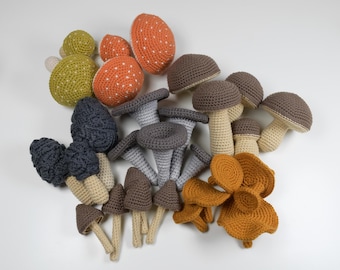 May include: A collection of crocheted mushrooms in various colors and styles, including red, orange, yellow, brown, and gray. The mushrooms are arranged in a cluster on a white background.
