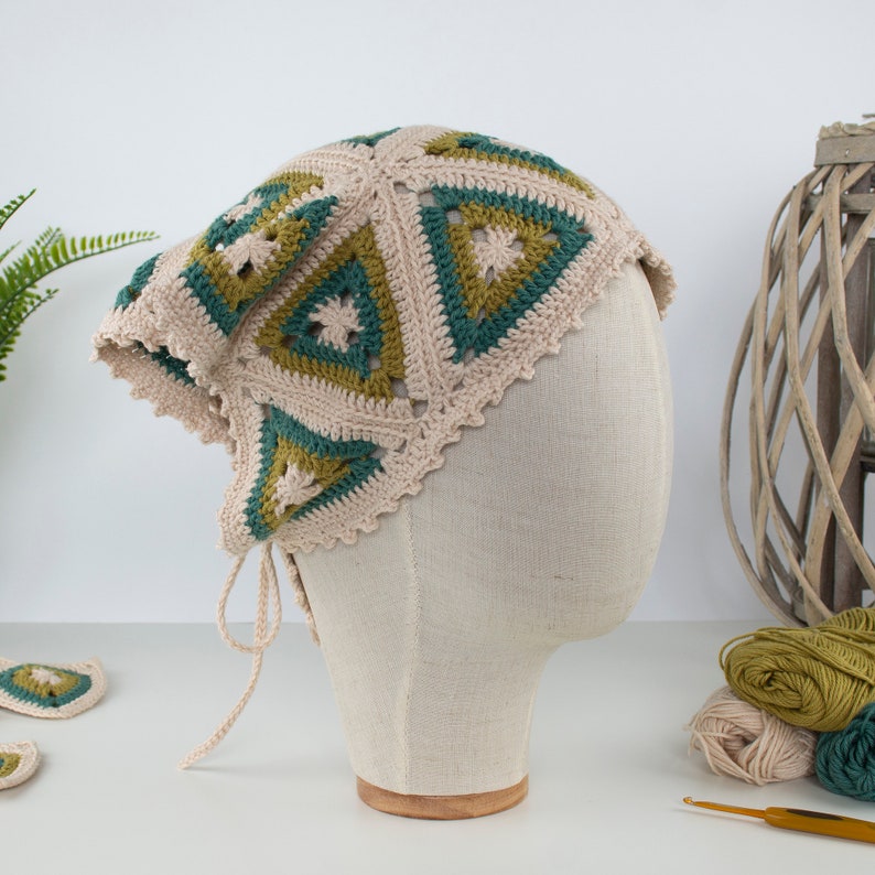 May include: A beige, green, and olive green crocheted hat with a triangular pattern. The hat has a scalloped edge and ties. The hat is displayed on a mannequin head.