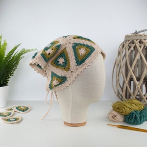 May include: A beige crochet head covering with green and olive-green triangle patterns. The head covering is displayed on a mannequin head. Crochet yarn and a crochet hook are visible in the background. The head covering has a scalloped edge and ties.
