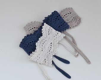 May include: Four crocheted bonnets in shades of gray, navy blue, and white are arranged on a white surface. Each bonnet features a scalloped edge and delicate openwork. The bonnets have long, knitted ties for securing under the chin. The bonnets are handmade.