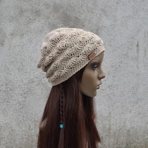 Op de afbeelding: Een beige gehaakte muts met een gestructureerd waaiermotief en een klein rechthoekig bruin leren label aan de zijkant. De muts heeft een slappe pasvorm en is gemaakt van lichtbruin garen, met gedetailleerd handwerk en een consistent steekpatroon.