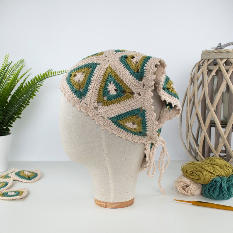 May include: A beige crochet head covering with green and teal triangle patterns. The head covering is displayed on a mannequin head. Beige yarn and a crochet hook are visible in the background.