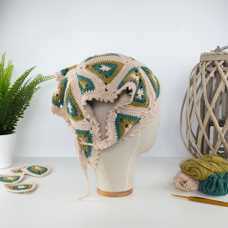 May include: A beige crochet hat with green and teal geometric patterns, displayed on a mannequin head. The hat has a scalloped edge and a tie. Several matching crochet pieces are visible, along with yarn and a crochet hook.