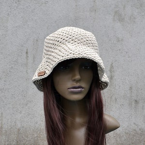 May include: A white crocheted bucket hat with a brown label. The hat is on a mannequin with long, dark brown hair.