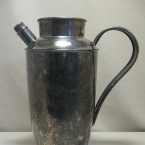 Op de afbeelding: Een zilveren cocktailshaker met een tuit en een handvat. De shaker heeft een aangetast oppervlak.