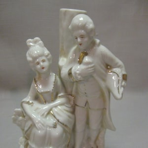 May include: A white porcelain figurine of a couple standing together. The woman is seated and the man is standing. Both figures are wearing 18th-century clothing and have gold accents.