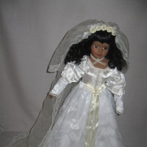 May include: A porcelain doll wearing a white wedding dress with a lace trim and a long veil. The doll has dark brown hair and brown eyes. She is wearing a pearl necklace and a white flower crown.