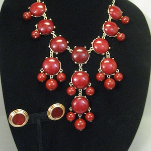 Puede incluir: Un collar de declaración rojo y dorado con grandes cuentas redondas rojas y cuentas rojas más pequeñas que cuelgan de la parte inferior. También se incluyen pendientes a juego rojos y dorados.
