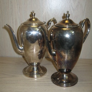 May include: Two silver teapots with ornate handles and lids. The teapots are on a wooden surface.