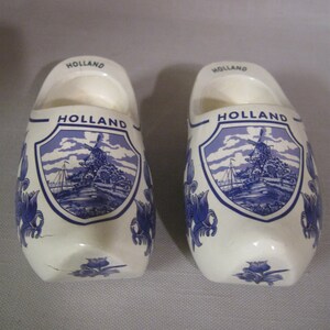 May include: Pair of white ceramic Dutch clogs with blue and white Delftware designs. Each clog features a windmill scene and the word "HOLLAND" written on the front and top.