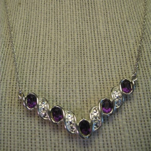 May include: A silver necklace with a curved design featuring five purple gemstones. The necklace is on a white fabric background.