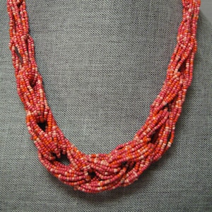Puede incluir: Un collar de múltiples hilos hecho de cuentas de semillas rosas y naranjas. El collar está anudado y retorcido, creando un diseño único e intrincado.