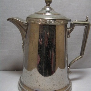 Puede incluir: Una cafetera vintage de color plateado con tapa y asa. La cafetera tiene un pico para verter y una banda decorativa alrededor de la tapa. El asa es rectangular y está unida al lateral. La cafetera es probablemente de metal.