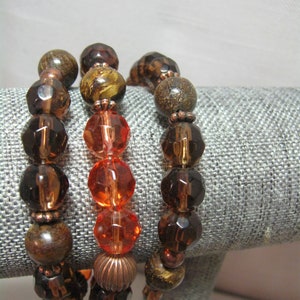 May include: Three bracelets with brown and orange beads. The bracelets are stacked on a gray fabric surface.