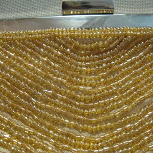 Puede incluir: Primer plano de un bolso de mano de cuentas doradas. El bolso est&aacute; hecho de peque&ntilde;as cuentas redondas que est&aacute;n cosidas juntas en un patr&oacute;n. Las cuentas son de color dorado claro y tienen un ligero brillo.