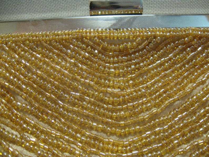 Puede incluir: Primer plano de un bolso de mano de cuentas doradas. El bolso est&aacute; hecho de peque&ntilde;as cuentas redondas que est&aacute;n cosidas juntas en un patr&oacute;n. Las cuentas son de color dorado claro y tienen un ligero brillo.