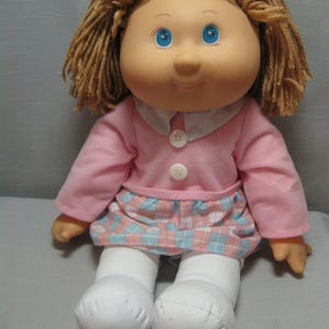 May include: A plush doll with a pink dress, white tights, and brown yarn hair. The doll has a smiling face with blue eyes.