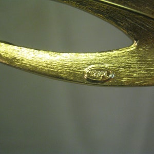 May include: Gold-toned metal brooch with a curved design and a 'TRIFARI' signature.