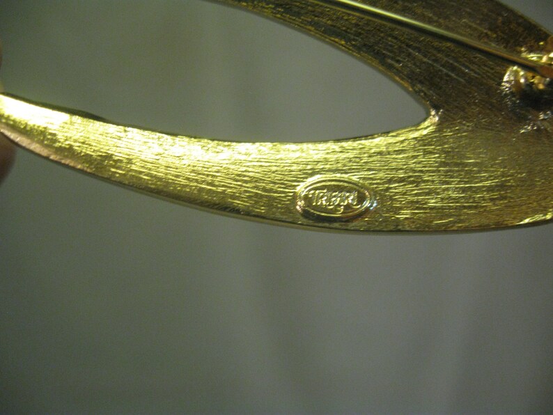 May include: Gold-toned metal brooch with a curved design and a 'TRIFARI' signature.