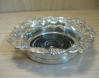 May include: Silver-plated wine bottle coaster with a decorative grape design. The coaster has a black rubber base.
