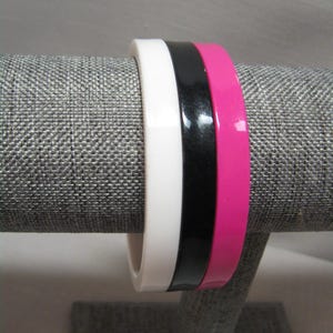 Puede incluir: Tres pulseras apiladas en blanco, negro y rosa intenso. Las pulseras están hechas de un material brillante y se exhiben en un soporte cubierto de tela gris. Las pulseras son un accesorio de moda atrevido.