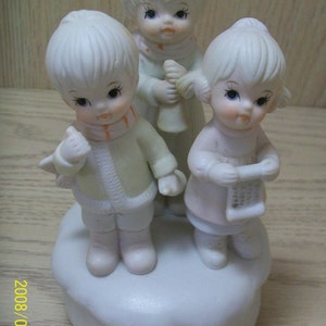 Könnte beinhalten: Eine Porzellanfigur-Gruppe mit drei Kindern, die Winterkleidung tragen. Die Kinder halten Musikinstrumente und stehen auf einem weißen Sockel. Die Figur ist eine Spieluhr.