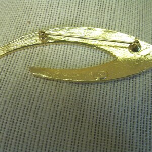 May include: Gold-toned metal brooch shaped like a crescent moon with a textured surface. The back of the brooch has a small, round, metal stamp with the text "Made in Italy".