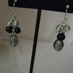 May include: A pair of dangle earrings with a cluster of beads. The earrings feature a silver hook and are adorned with a mix of gray and black beads. The beads vary in shape and size, creating a layered effect.