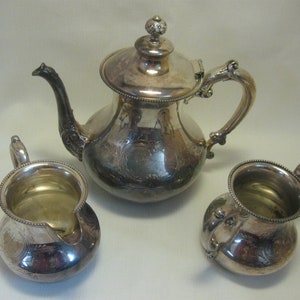 May include: A silver tea set, including a teapot with a lid and two small creamers. The set has ornate handles and decorative floral patterns. The teapot has a curved spout and a decorative finial on the lid.