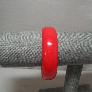 Puede incluir: Un brazalete rojo brillante. El brazalete es de color rojo liso y brillante y parece estar hecho de un material liso. El brazalete se exhibe en un expositor de joyería gris. El diámetro del brazalete es de aproximadamente 6,35 cm.