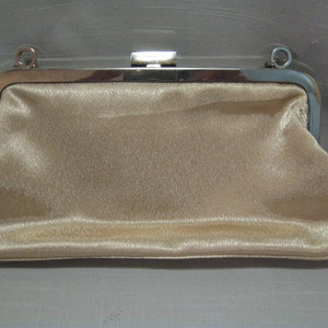 Puede incluir: Un bolso de mano de sat&eacute;n beige con un marco de metal plateado y una cadena.