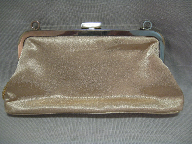 Puede incluir: Un bolso de mano de sat&eacute;n beige con un marco de metal plateado y una cadena.