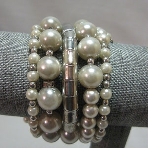 Può includere: Un set di quattro braccialetti di perle con accenti in metallo argentato. I braccialetti sono impilati su un espositore in tessuto grigio.