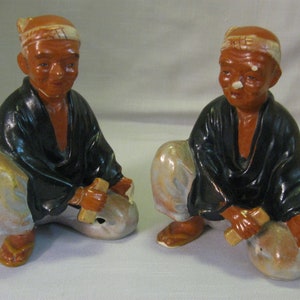 Op de afbeelding: Twee keramische beeldjes van mannen in traditionele Japanse kleding. De mannen zitten met gekruiste benen en houden kleine houten gereedschappen vast. De beeldjes zijn geschilderd in tinten bruin, zwart en wit.