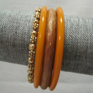 May include: A collection of bracelets in shades of gold, amber, and brown. The bracelets include a gold beaded bracelet, two amber-colored bangles, and a brown, textured bangle. The bracelets are displayed on a gray surface.