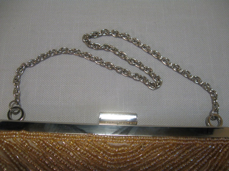 Puede incluir: Un peque&ntilde;o bolso de noche de cuentas con una correa de cadena plateada. El bolso est&aacute; hecho de una tela marr&oacute;n claro y tiene un cierre plateado.