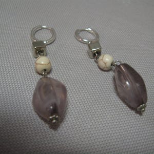 May include: A pair of dangle earrings featuring silver-tone hoops, square accents, and cream-colored beads. Each earring has a large, oval, translucent, lavender-colored bead with a silver-tone cap.