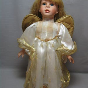 Pode incluir: Uma boneca de porcelana vestida de anjo com cabelo loiro, olhos azuis e um vestido branco com detalhes dourados. A boneca usa uma auréola dourada e tem asas douradas.