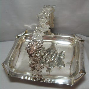 May include: Silver serving tray with a grapevine handle. The tray is square with rounded corners and has a decorative border. The handle is made of a single piece of metal that is shaped like a grapevine with leaves and grapes.