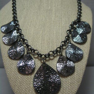 May include: A silver-toned necklace with a textured finish. The necklace features a chain with multiple teardrop-shaped pendants of varying sizes. The pendants have a hammered metal appearance, creating a unique, eye-catching design. The necklace is displayed on a neutral-colored bust.