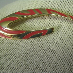 May include: A gold-toned brooch with a red and gold abstract design. The brooch is shaped like a crescent moon.