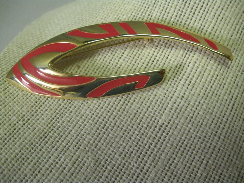 May include: A gold-toned brooch with a red and gold abstract design. The brooch is shaped like a crescent moon.