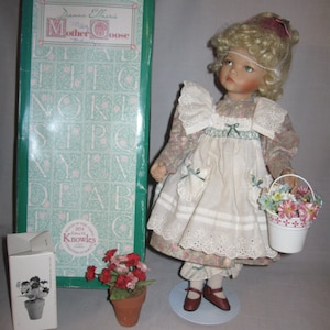 Puede incluir: Una muñeca de porcelana con cabello rubio rizado que lleva un vestido floral y un delantal blanco. Ella sostiene un cubo blanco con flores rosas y amarillas. La muñeca está de pie sobre un soporte blanco. La muñeca es parte de la colección 'My Mother Goose' de Donna Ethier.