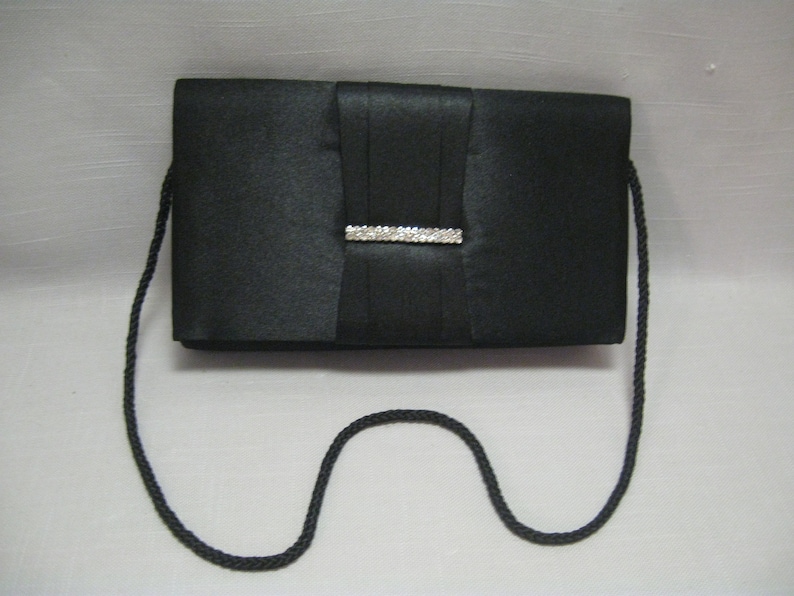 May include: A black satin clutch bag with a rhinestone embellishment and a long black strap.