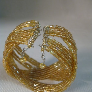 Puede incluir: Un brazalete de cuentas doradas con m&uacute;ltiples hileras de cuentas y abalorios colgantes. El brazalete est&aacute; hecho de peque&ntilde;as cuentas redondas que est&aacute;n entrelazadas en un patr&oacute;n intrincado.
