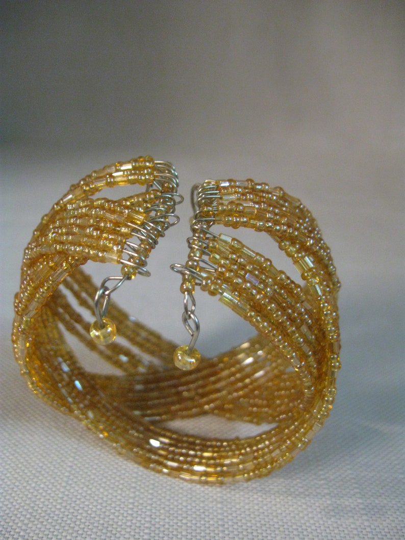 Puede incluir: Un brazalete de cuentas doradas con m&uacute;ltiples hileras de cuentas y abalorios colgantes. El brazalete est&aacute; hecho de peque&ntilde;as cuentas redondas que est&aacute;n entrelazadas en un patr&oacute;n intrincado.