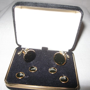 May include: A set of cufflinks and shirt studs in a black velvet box. The cufflinks and studs have a gold-tone border and a black, circular center. The box has a black exterior and a white interior.