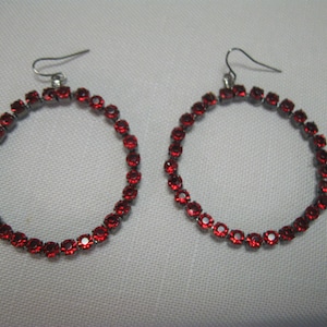 Puede incluir: Un par de pendientes de aro con una montura de metal plateado. Cada pendiente está adornado con numerosas piedras preciosas rojas pequeñas y redondas, creando un círculo continuo. Los pendientes tienen un cierre de gancho simple.