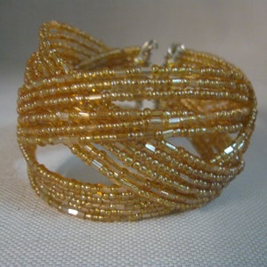 Puede incluir: Un brazalete de cuentas doradas con un dise&ntilde;o tejido. El brazalete est&aacute; hecho de peque&ntilde;as cuentas redondas que est&aacute;n enhebradas juntas en un patr&oacute;n de cruz.