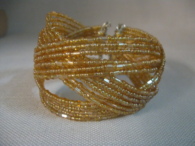 Puede incluir: Un brazalete de cuentas doradas con un dise&ntilde;o tejido. El brazalete est&aacute; hecho de peque&ntilde;as cuentas redondas que est&aacute;n enhebradas juntas en un patr&oacute;n de cruz.